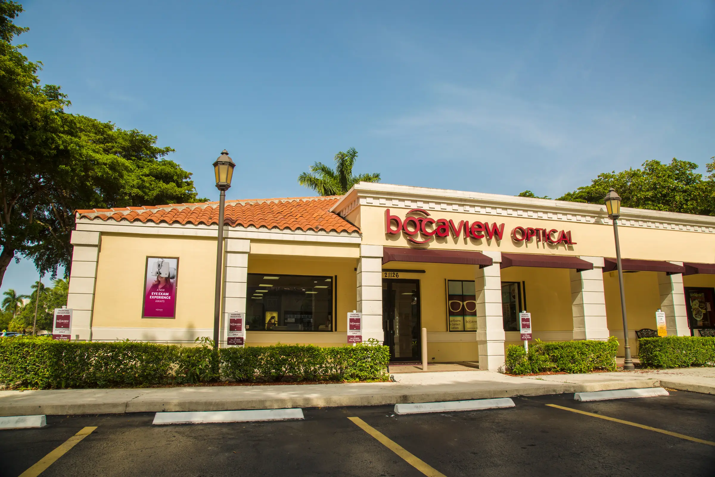 Single-story building of BocaView Optical with a tan exterior and red signage. Surrounded by greenery and a clear blue sky, it has parking spaces in front.